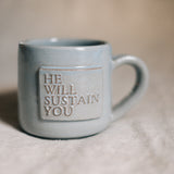 Blue ceramic mug with 'HE WILL SUSTAIN YOU' text on a beige surface