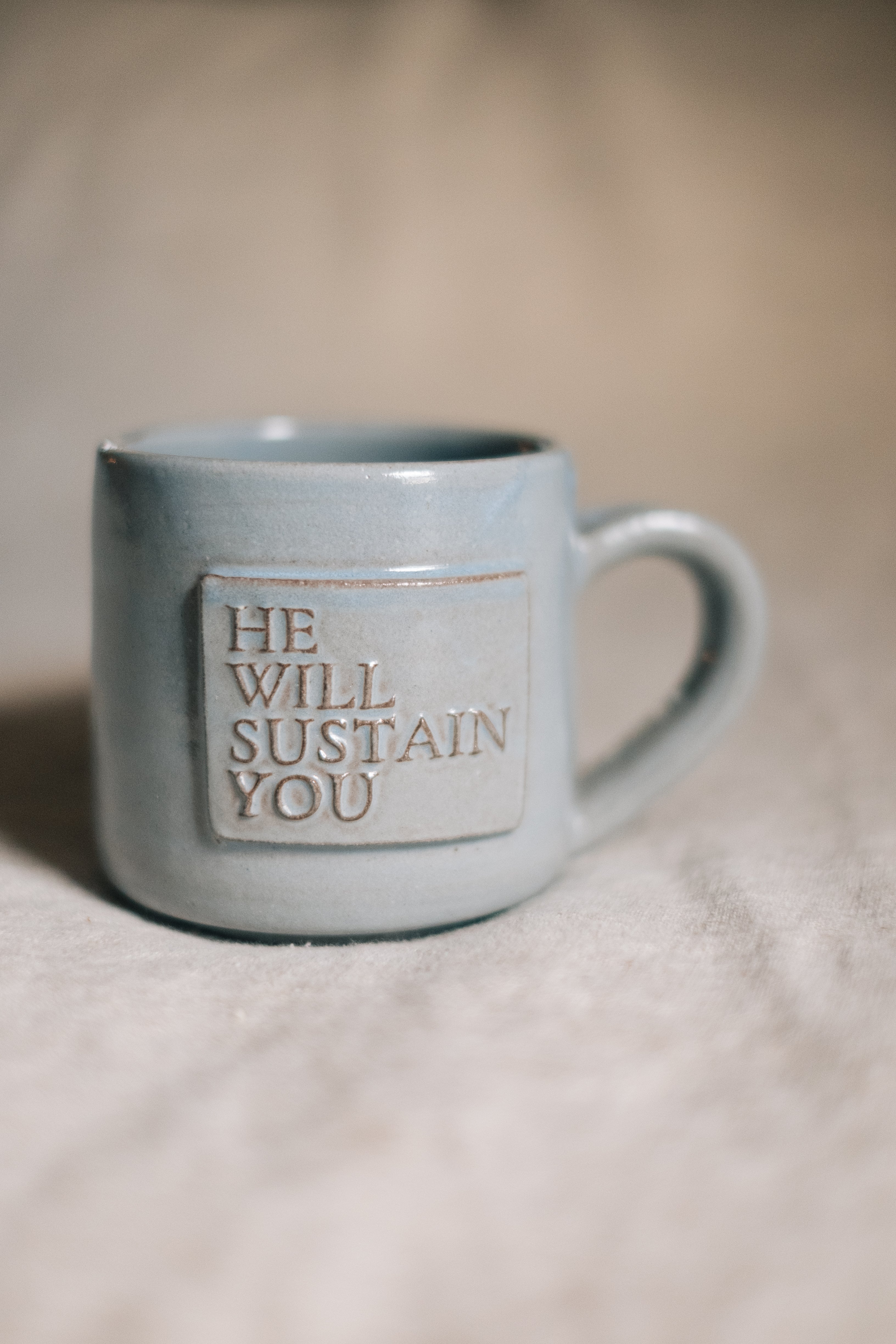 Blue ceramic mug with 'HE WILL SUSTAIN YOU' text on a beige surface