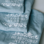 Three folded dusty blue graphic t-shirts stacked diagonally, each printed with the phrase 'HE RESTORES MY SOUL' in white serif lettering, surrounded by delicate white floral illustrations.
