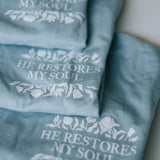 Three folded dusty blue graphic t-shirts stacked diagonally, each printed with the phrase 'HE RESTORES MY SOUL' in white serif lettering, surrounded by delicate white floral illustrations.