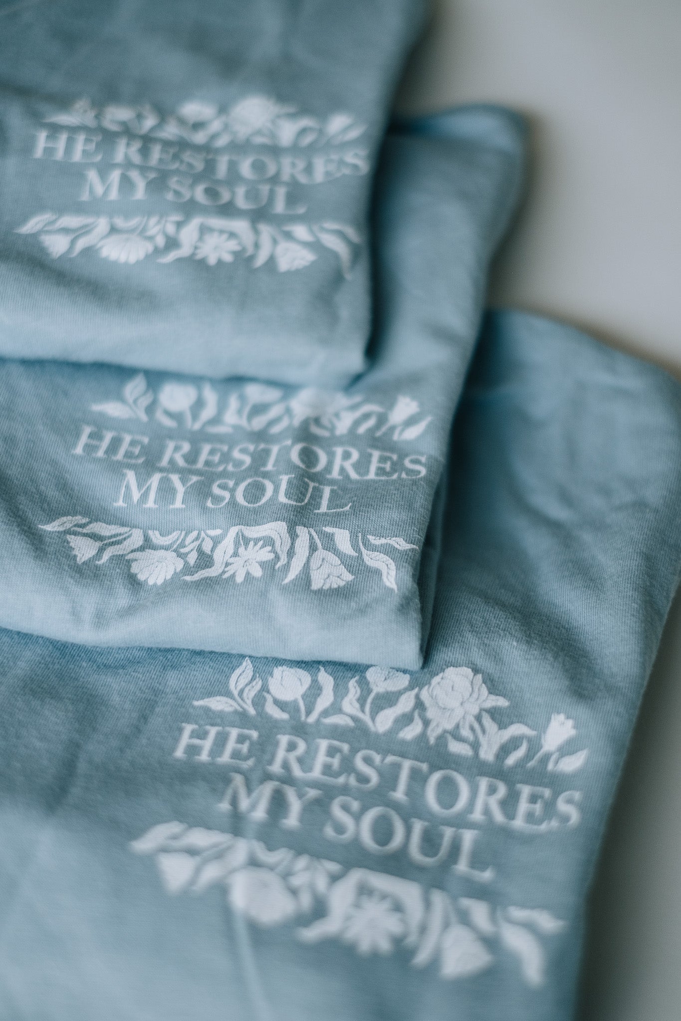 Three folded dusty blue graphic t-shirts stacked diagonally, each printed with the phrase 'HE RESTORES MY SOUL' in white serif lettering, surrounded by delicate white floral illustrations.