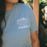 A woman wearing a light blue t-shirt with a white graphic that reads 'HE RESTORES MY SOUL' surrounded by floral illustrations, set against a blurred outdoor background.