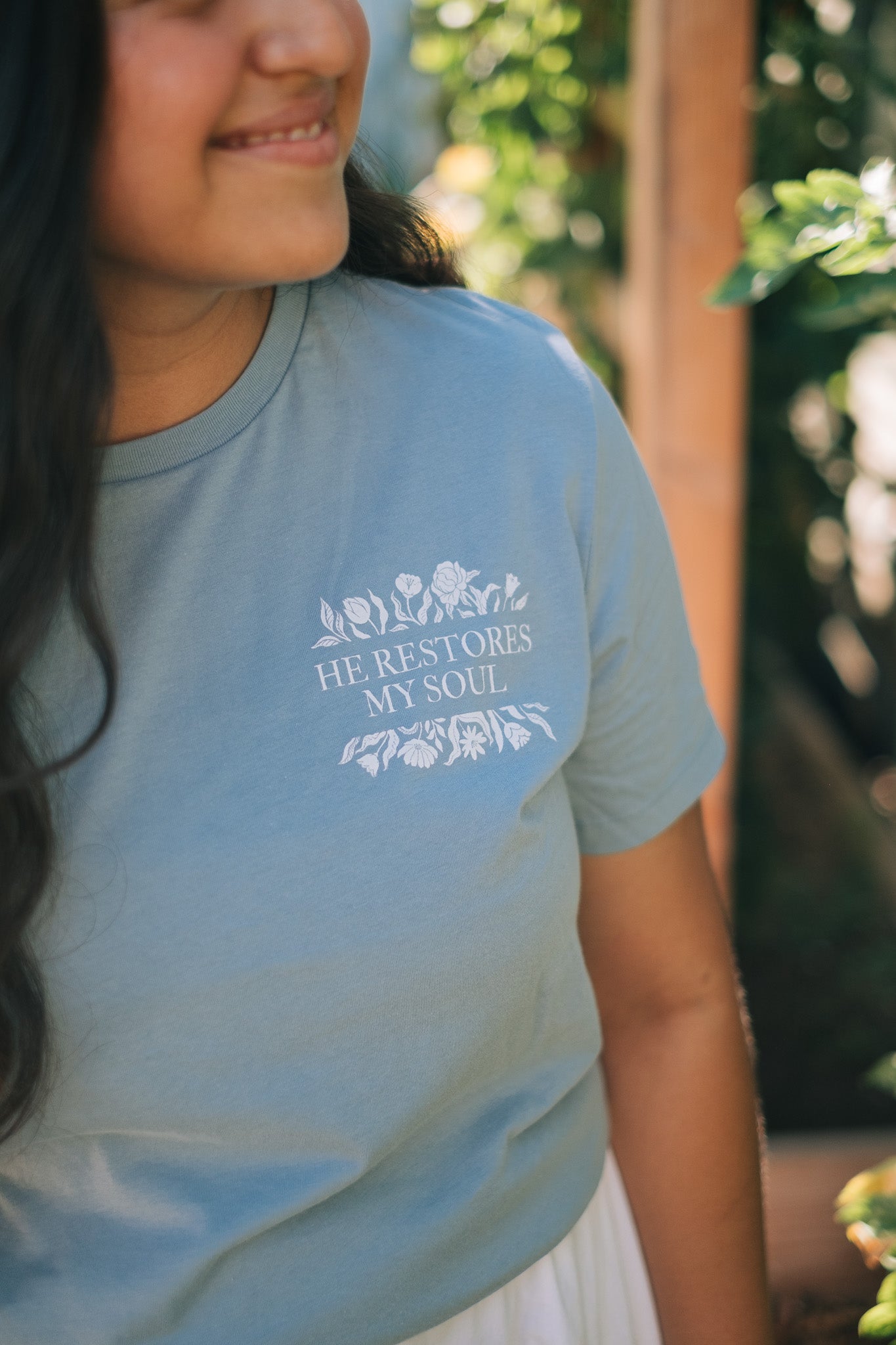 A woman wearing a light blue t-shirt with a white graphic that reads 'HE RESTORES MY SOUL' surrounded by floral illustrations, set against a blurred outdoor background.