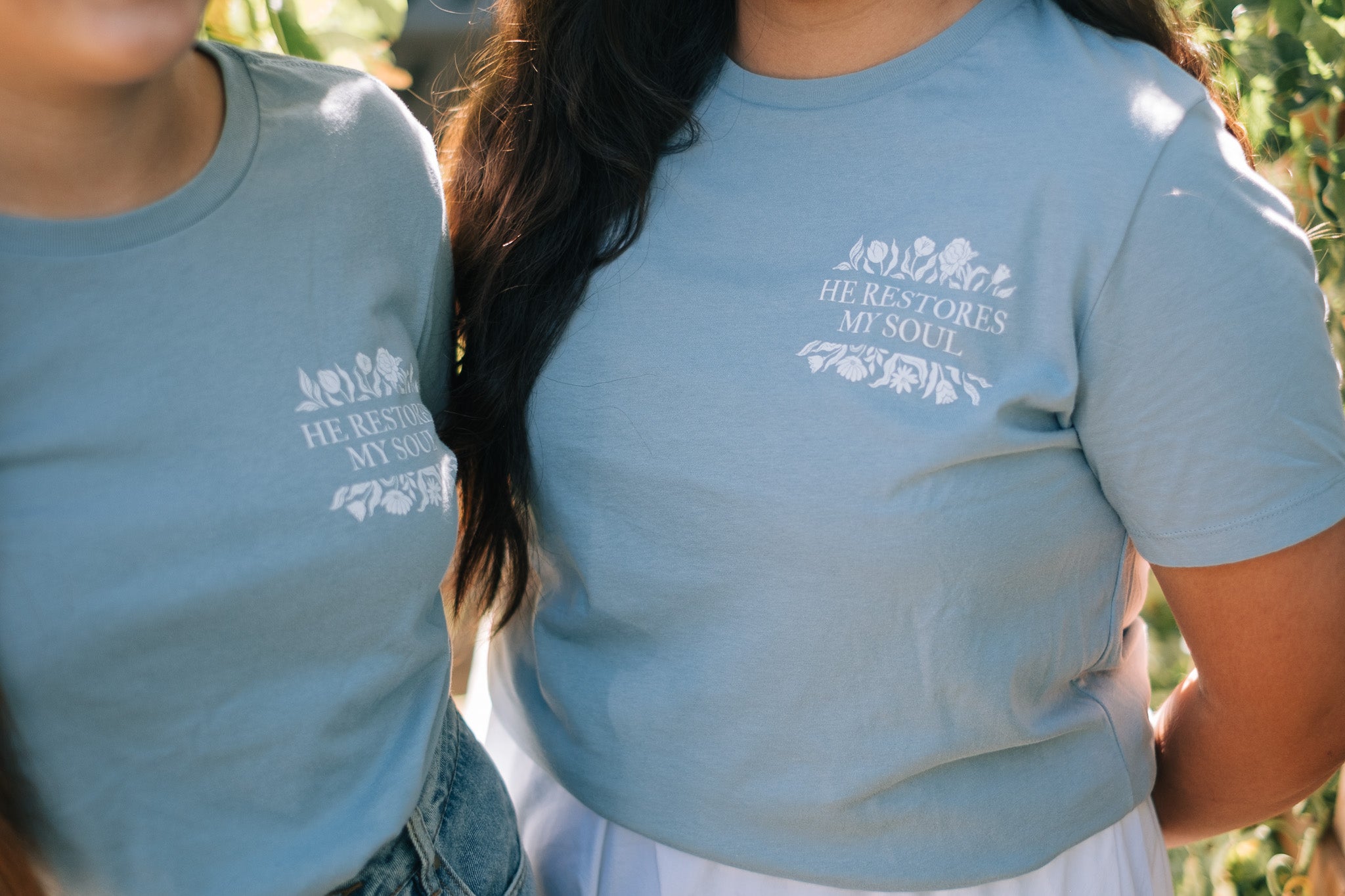 A close-up lifestyle shot of two people in a garden wearing matching light blue t-shirts with a white graphic print that reads 'HE RESTORES MY SOUL' with a small floral illustration.