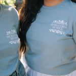 A close-up lifestyle shot of two people in a garden wearing matching light blue t-shirts with a white graphic print that reads 'HE RESTORES MY SOUL' with a small floral illustration.