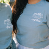 A close-up lifestyle shot of two people in a garden wearing matching light blue t-shirts with a white graphic print that reads 'HE RESTORES MY SOUL' with a small floral illustration.