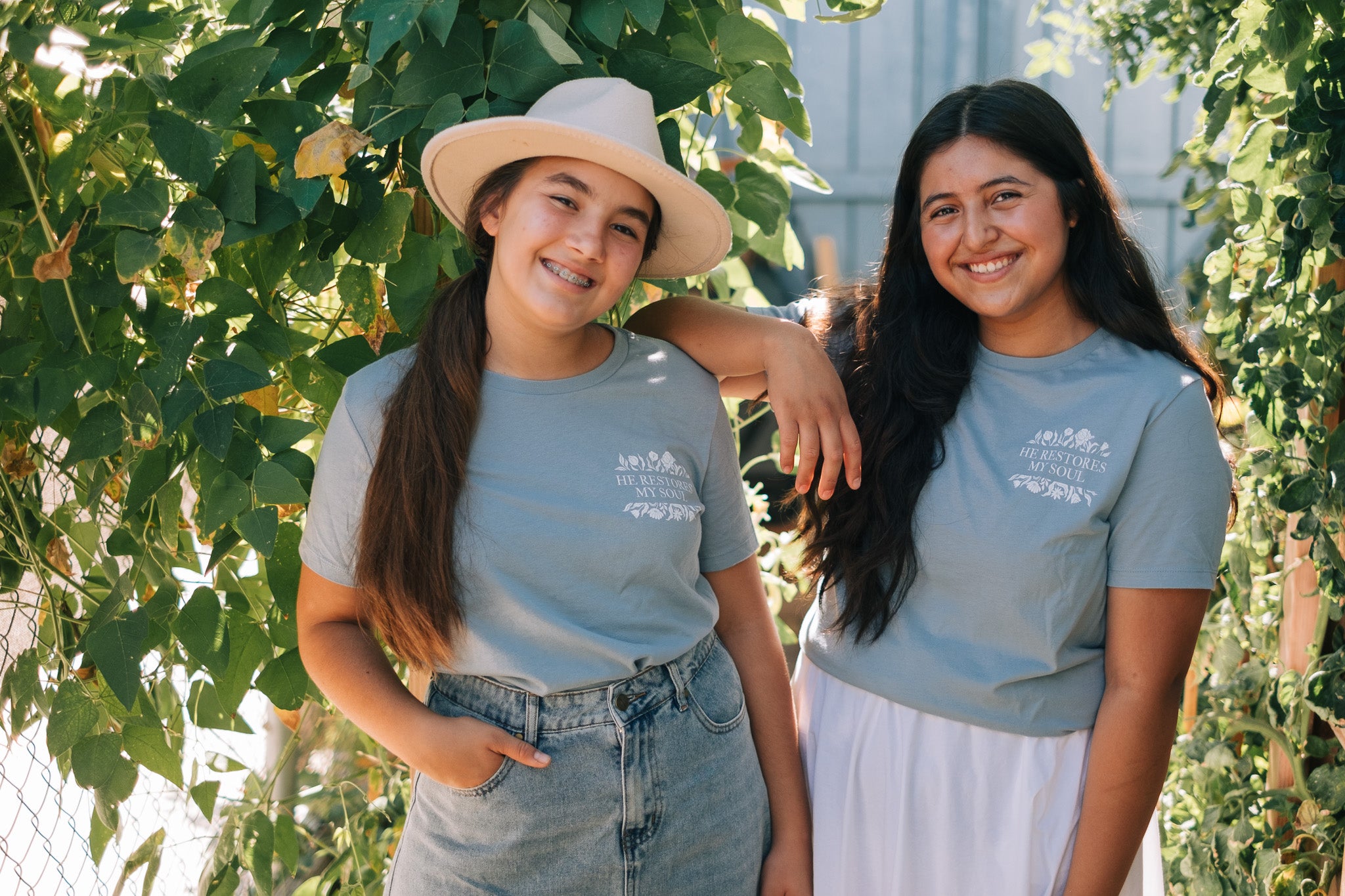 Two smiling women standing outdoors among leafy green vines, wearing matching light blue-gray T-shirts with a small white graphic on the chest and light-wash denim bottoms.
