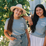 Two smiling women standing outdoors among leafy green vines, wearing matching light blue-gray T-shirts with a small white graphic on the chest and light-wash denim bottoms.