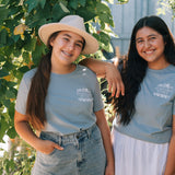 Two smiling women standing outdoors among leafy green vines, wearing matching light blue-gray T-shirts with a small white graphic on the chest and light-wash denim bottoms.