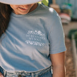A young lady wearing a light blue graphic t-shirt with the text 'HE RESTORES MY SOUL' and a floral design, paired with a white wide-brim hat and denim jeans in an outdoor setting.