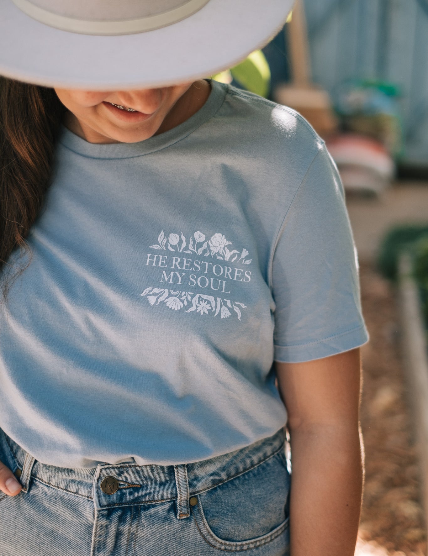A young lady wearing a light blue graphic t-shirt with the text 'HE RESTORES MY SOUL' and a floral design, paired with a white wide-brim hat and denim jeans in an outdoor setting.