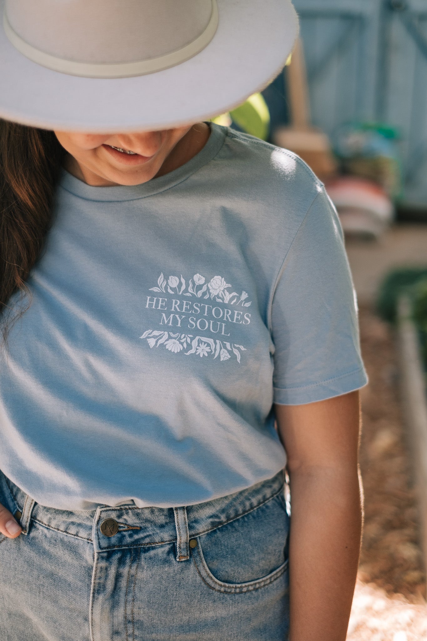 A young lady wearing a light blue graphic t-shirt with the text 'HE RESTORES MY SOUL' and a floral design, paired with a white wide-brim hat and denim jeans in an outdoor setting.