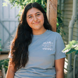 A woman with long dark hair stands outdoors in a garden, wearing a light blue crewneck t-shirt with a white graphic print on the left chest that reads 'HE RESTORES MY SOUL'.