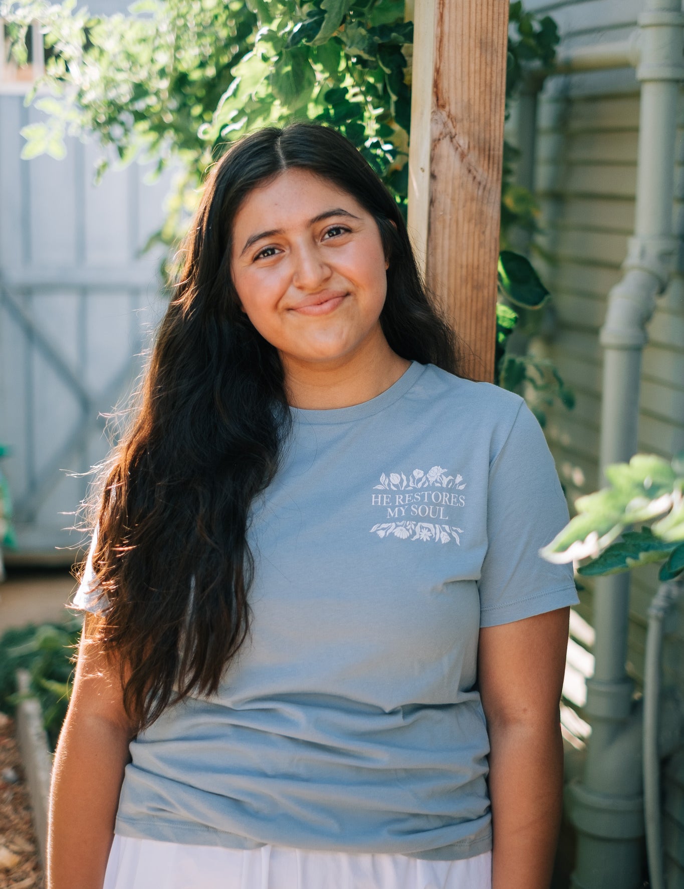 A woman with long dark hair stands outdoors in a garden, wearing a light blue crewneck t-shirt with a white graphic print on the left chest that reads 'HE RESTORES MY SOUL'.
