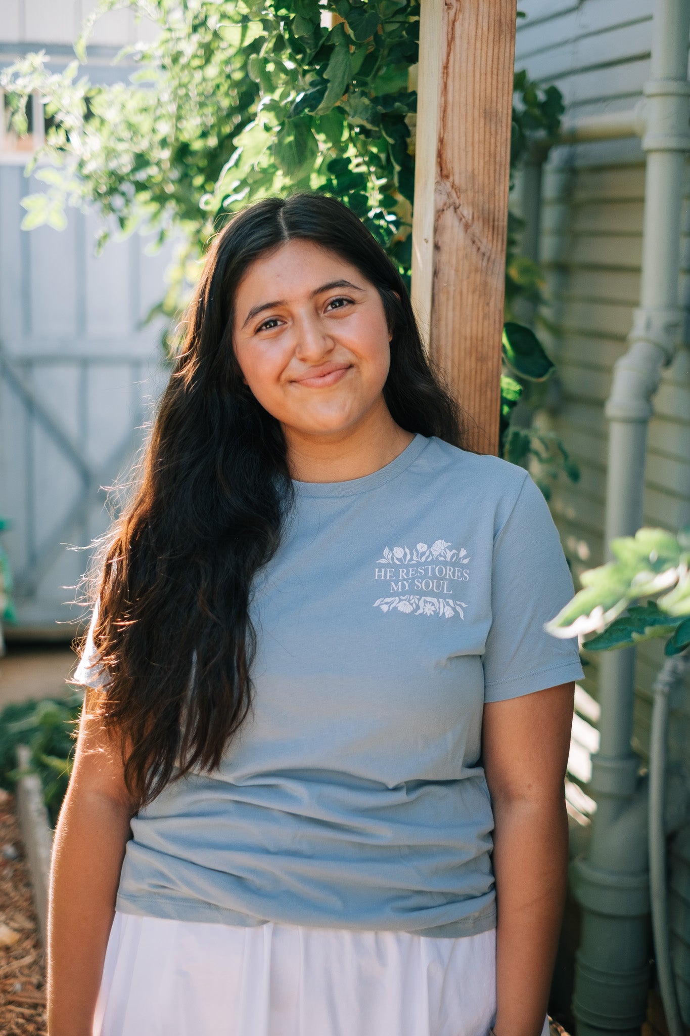 A woman with long dark hair stands outdoors in a garden, wearing a light blue crewneck t-shirt with a white graphic print on the left chest that reads 'HE RESTORES MY SOUL'.